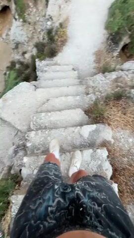 A first-person perspective video shows a person walking down a stone staircase. The person is wearing black and blue patterned shorts and white sneakers. They are on a path next to a rocky cliff overlooking the ocean. The path leads to a cave entrance in the cliff face. The video pans to show the ocean, cliffs, and the cave.