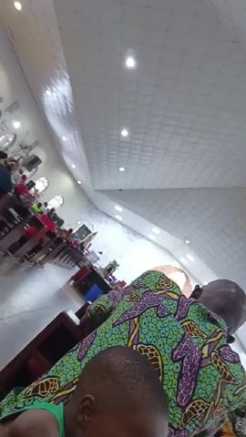 The video shows the interior of a church or religious building during what appears to be a worship service. The camera is positioned low, looking upwards and forwards from behind a few congregants. In the immediate foreground, the back of a young child's head, wearing a green top, is visible. Behind the child, the back of an adult male's head and shoulders are seen. He is wearing a vibrantly patterned short-sleeved shirt, predominantly green with purple, yellow, and possibly black or dark blue abstract or animal-like motifs.

The ceiling of the building is white with a distinctive quilted or grid-like pattern and features several recessed circular lights that illuminate the space. Further back on the left side of the frame, rows of wooden pews are visible, occupied by numerous other congregants. Some individuals in the pews appear to be wearing red shirts. In the far background, towards the front of the church, there's a glimpse of the altar area, though it's not clearly defined due to the perspective. The overall lighting is bright and even. The sound of singing is audible throughout the video, indicating a communal worship activity.