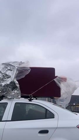 The video shows a white car driving on a highway on a cloudy day. The car has a roof rack with various items strapped to it, including a maroon cushion, items wrapped in foil, and plastic sheeting. The camera appears to be inside another vehicle moving alongside the white car. The roadside features a metal guardrail and a modern building with large windows. The road is wet.