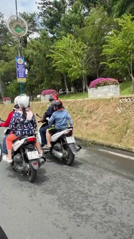 Two motorcycles are traveling on a paved road. The first motorcycle has three people on it: a person driving, and two passengers behind. The second motorcycle has two people on it: a person driving, and one passenger behind. All individuals are wearing helmets. The road is lined with trees and a grassy embankment. A signpost with writing and a decorative element is visible on the embankment. The sky is cloudy. The motorcycles appear to be moving forward.