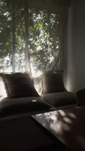 Here's a concise description of the video:

The video shows a sun-drenched window seat with two large, square pillows. The pillows are patterned and have a light fringe. A sheer, white curtain hangs in front of the window, creating dappled shadows on the pillows and the nearby wall. Outside the window, green foliage is visible. In the foreground, a corner of a wooden table is partially visible, also covered in shadows. The scene is bathed in warm, natural light.
