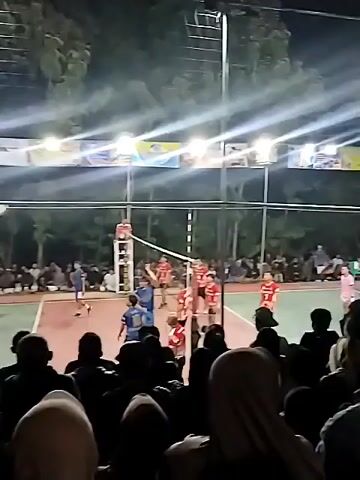 A volleyball game is being played at night under floodlights. There is a net dividing the court, and players in red and blue uniforms are on opposing sides. Spectators are gathered around the court, some sitting and some standing. The atmosphere appears to be energetic and engaged.