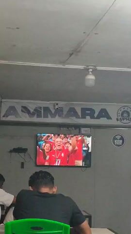 The video captures a scene inside a dimly lit room, possibly a cafe or waiting area. In the foreground, a person with dark hair is seen from behind, seated at a table and facing away from the camera. They are wearing a black t-shirt. In the mid-ground, a large flat-screen television is mounted on a gray wall, displaying a broadcast of a sporting event. The screen shows a crowd of people, mostly wearing red, cheering. Above the television, a banner with the word "AMMARA" in large, dark blue letters is prominently displayed. To the right of the banner, a circular logo with the text "KUPH" is visible. A small circular sticker with a Wi-Fi symbol and the words "WIFI FREE" is also affixed to the wall near the television. The ceiling is made of textured material, with a light fixture protruding from it. The overall impression is one of casual viewing in a public or semi-public space.