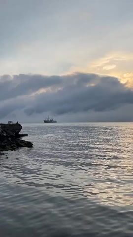 The video shows a body of water with a ship in the distance. The sky is overcast with some lighter clouds showing the sun. There is a rocky shore in the foreground with some greenery. The water is calm and reflects the sky.