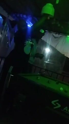 Two people are playing pool on a green pool table with several balls on it. The pool table is black with a logo on the side. The location is poorly lit but overhead lighting can be seen. The people are standing behind a low metal fence covered by clear plastic. One person is wearing a black shirt with a colorful sun on it and dark pants with a single stripe down the side. The other person is wearing a light gray sweatshirt with a dark collar and dark shorts.