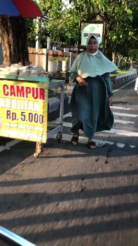 A woman wearing a light green hijab and a dark green dress walks across a crosswalk. She is carrying a bag in her left hand. To her left, a food cart with a sign that reads "CAMPUR KHOLIFAH RP. 5.000" is visible. The cart is filled with food items. In the background, there is a street with traffic markings, a sidewalk, trees, and a sign for "SDN LARANGAN". The lighting suggests it is daytime, possibly late afternoon due to the long shadows. The ground is paved with asphalt and has painted white lines for the crosswalk.