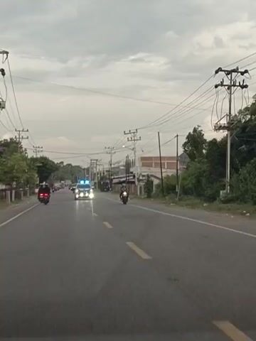 The video shows a highway with traffic. On the road, there are several vehicles including motorcycles, a police car with flashing lights, and other cars. The sky is overcast, and there are power lines running overhead. The environment appears to be semi-urban or rural with buildings and trees visible on the sides of the road. The video captures the movement of vehicles on the road, suggesting traffic flow or possibly a convoy.