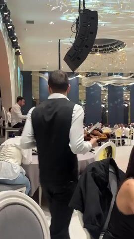 The video shows the interior of what appears to be a large, upscale event space, possibly a ballroom. The scene is set with dining tables and chairs, some of which are occupied by guests. A band consisting of at least three members—a keyboardist, a clarinetist, and another musician—is playing live music. The band is positioned on a small stage area. A waiter, dressed in a white shirt and black vest, walks through the room carrying a tray with what appears to be food. Overhead, there are large speaker arrays suspended from the ceiling and decorative light fixtures.
