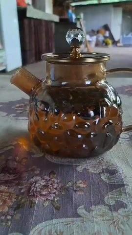 A glass teapot is tilted to pour a dark amber liquid into a clear glass mug decorated with cartoon characters. The teapot is ornate, with a textured glass body and gold accents, topped with a crystal lid. The pouring action begins at the 00:04 mark.