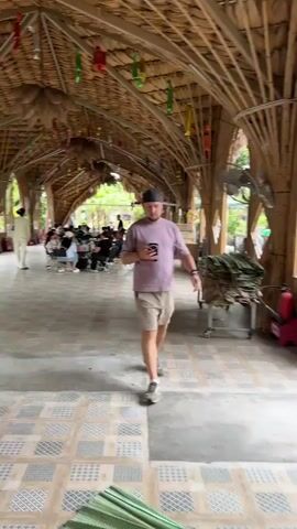 A man wearing a light purple t-shirt and khaki shorts is walking towards the camera holding a smartphone. He is in a covered outdoor seating area with a thatched roof supported by bamboo poles. The floor is tiled with a patterned design. In the background, several tables and chairs are set up, with some people seated at them. There are decorative items hanging from the ceiling. To the right, a cart with folded green mats and other items is visible, along with a red fire extinguisher. A long wooden bench is in the foreground to the left. The overall environment appears to be a casual, possibly tropical, dining or resting area.