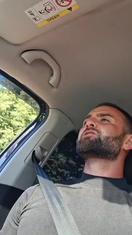 A man with a beard is in the driver's seat of a car. He is wearing a grey shirt and a seatbelt. He is looking up and to his left, speaking. The sky is visible through the car window. There is a handle on the roof of the car, and an airbag warning sticker.