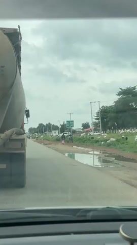 This video captures a roadside scene in Kwali, Nigeria, under a cloudy sky, suggesting it's daytime with overcast weather. The perspective is from inside a vehicle, looking out through the windshield.

The primary subjects are several cows grazing peacefully on the side of a paved road. The road itself appears to have puddles and some disrepair, indicating potential recent rainfall or poor drainage. In the distance, a large truck is visible on the left, suggesting ongoing traffic.

Further down the road, a few people are walking. One individual, dressed in a pink garment, is prominent. A green road sign is also clearly visible, providing directional information. The sign indicates distances to various locations: Zaria (284km), Kaduna (159km), and Abuja (34km), confirming the location as being on a major route in Nigeria.

The overall scene depicts a typical rural or semi-urban roadside environment in Nigeria, characterized by the presence of livestock, road infrastructure, and people going about their day. The cloudy weather contributes to a muted visual palette, and the presence of puddles suggests a humid or rainy climate. There are no overt signs of distress or urgency; the atmosphere appears calm.
