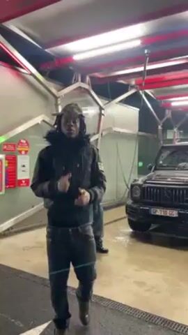 Here is a description of the video.
The video appears to be a music video of an individual dancing and singing in front of a car wash with a black Mercedes-Benz G-Wagon. The main individual is wearing a dark puffy coat, grey jeans and a cap with a colorful pattern. Behind him in the background, another person is standing beside the G-Wagon. The person in the foreground dances and then points upwards towards the red car wash sign. The video then transitions to show the G-Wagon covered in soap. The scene suggests they are waiting for it to be cleaned. In the background, someone uses a brush to clean the car.