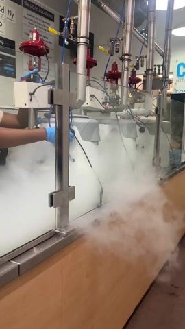 A static, medium shot captures the interior of what appears to be a food service establishment, possibly an ice cream shop. A person wearing blue gloves is visible behind a transparent, glass barrier. They are operating a machine with a "Globe" logo, which is generating a significant amount of white, cryogenic fog. Above the machine, there are pipes and red valves. A laminated sign is to the left, displaying menu options. The counter is made of wood. Behind the machine, a sign is partially visible saying "Chi ... NITRO".