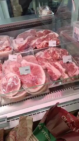 The video displays a refrigerated meat counter in a grocery store or butcher shop, likely located in Kuleshovka, Russia. The glass-fronted display case is brightly lit with a cool, slightly pinkish light, illuminating various cuts of raw meat.

The shelves are filled with pre-packaged portions of fresh meat, predominantly pork, judging by the color and fat marbling. Each cut is wrapped in clear plastic film or bags, and many are placed on white foam trays. Visible cuts include large, thick pieces that appear to be pork shoulder or roasts, as well as thinner slices resembling pork chops or steaks, and some cuts with visible bone and fat, possibly ribs or belly meat.

Each packaged item has a white rectangular price tag with red numbers and the Russian Ruble symbol (₽). Clearly visible prices include "395₽", "365₽", "405₽", "615₽" (partially obscured), "355₽", and "325₽".

In the bottom right corner of the frame, a brown paper bag with green and white details is partially visible. Cyrillic text on the bag includes "УГОЛЬ" (Ugol), which translates to charcoal, suggesting it's a bag of charcoal, often sold in proximity to meat for grilling. Another patterned bag is partially visible to its left.

No people are explicitly visible in the frame, but the perspective suggests a shopper viewing or recording the display. The setting is clearly an indoor retail environment.