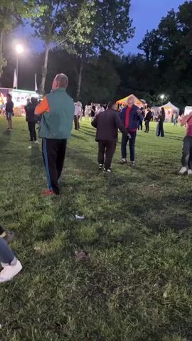 The video shows a group of people dancing on a grass field at what appears to be a fair or festival. There are various tents and structures set up in the background, including one that resembles a small carousel. People are milling around, some watching the dancers. Three men are prominent in the foreground: one in a green vest and Adidas pants, another in a dark jacket and pants, and a third in a pink shirt and green pants, who carries a black jacket. The scene is illuminated by overhead lights and the time appears to be dusk or early evening. The field is littered with some trash.