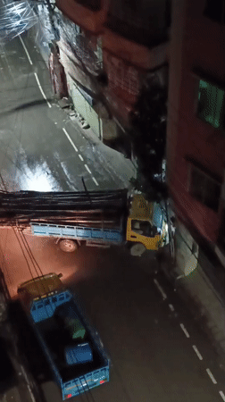 Rainy night on a narrow Dhaka street, trucks parked, pedestrians passing