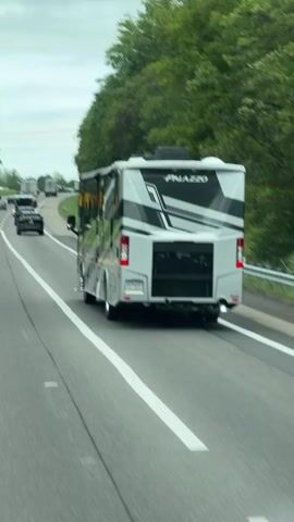 The video shows the rear view of a large, modern recreational vehicle (RV), identified by the name "PALAZZO" on its back. The RV is gray with black and white geometric accents. It is traveling on a highway, indicated by the white lines on the asphalt and the presence of other vehicles in the background. The RV is in the right lane, and there are several other vehicles in front of it, including cars and semi-trucks. The environment appears to be a rural area, with a dense line of green trees lining the right side of the highway. The sky is overcast with a pale, cloudy appearance. The camera is positioned behind and slightly to the left of the RV, capturing its movement down the highway.