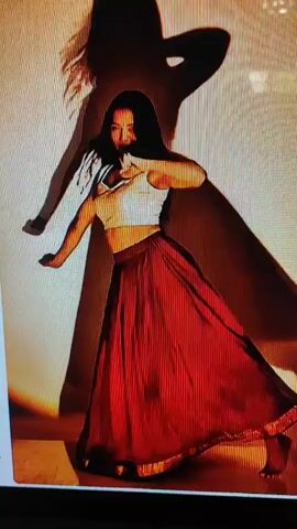 A woman in a white crop top and a red skirt dances in front of a wall. Her shadow is projected on the wall behind her. The lighting is warm and atmospheric, with a spotlight on the floor. The woman moves gracefully, her skirt swirling around her. She smiles and engages with the camera, her movements fluid and expressive.
