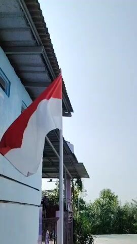 A flag with red and white horizontal stripes, likely the Indonesian flag, is visible on a flagpole. The flag is partially unfurled and appears to be waving in the wind. The background shows a light blue sky, the side of a building with a white and blue exterior, a corrugated roof, and trees in the distance. The image is captured from a low angle, looking up towards the flag.