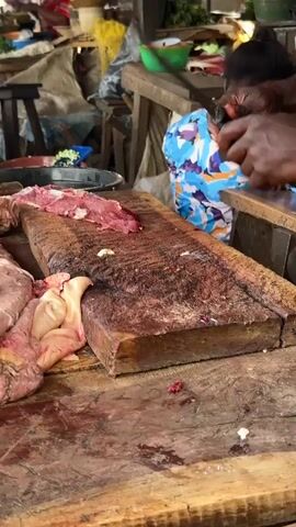 A close-up shot captures a butcher's workspace. A large wooden chopping block, stained with what appears to be blood and meat residue, dominates the foreground. Raw meat, some with visible fat and bone, is arranged on the block. A butcher, with dark skin and wearing a dark apron, is actively using a large cleaver to cut meat. In the background, another person, wearing a blue and white patterned garment, is partially visible, also seemingly engaged in food preparation. The environment suggests a bustling market or food stall, with glimpses of other stalls and produce in the blurred background. The lighting is dim, typical of an indoor or covered market setting.
