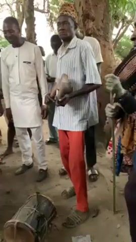 A group of men are gathered outdoors, seemingly for a ceremonial or cultural event. One man in the center wears a leopard print hat, a striped shirt, and red pants. He holds a bowl-like object and strikes it with a small wooden stick. To his left, a man in a white outfit stands by. Behind them, more people are gathered, some looking towards the man striking the bowl. On the ground, there's a drum lying next to the man in the red pants. To his right, another man is holding a cane and is wearing a brightly colored robe. They are standing on ground covered with dirt and some debris, under the shade of trees.