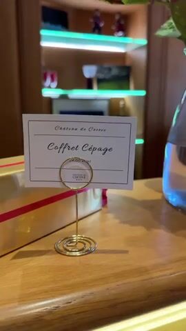 A close-up shot shows a wooden counter with a small card in a gold wire stand that reads "Château de Cassan Coffret Cépage." To the left is a gold gift box with pink ribbon. To the right, two black wine bottle-shaped cases are open, revealing wine accessories. The left case contains a wine stopper and aerator, while the right case holds a corkscrew. In the background, a dark-colored wine bottle stands on a shelf with LED lighting.
