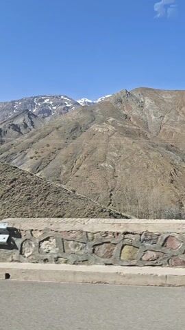 The video shows a view of mountains with snow-capped peaks against a clear blue sky. The camera appears to be positioned in a moving vehicle, looking out over a low stone wall and a silver guardrail. The mountains are rocky and covered with sparse vegetation. Power lines are visible on one of the slopes. The road is asphalt.