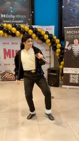A male performer dressed as Michael Jackson, wearing a black sequined jacket, white shirt, black pants, and black shoes with white socks, stands center frame in front of a balloon archway made of black and gold balloons. He holds a microphone in his right hand and gestures with his left arm. Behind him are posters for "MUVIX CINEMAS" and "MICHAEL JACKSON." To his left is a poster with "Dolby Atmos" and to his right is a large banner featuring an image of Michael Jackson. The floor is tiled. The background appears to be an indoor venue, possibly a cinema lobby. The performer is mid-performance, singing or speaking into the microphone.