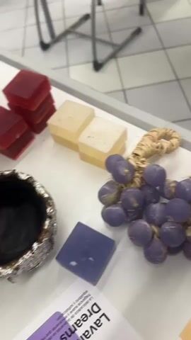 This is a close-up, slightly blurry video of a table displaying various handcrafted items, likely candles or soaps, with accompanying product cards. The setting appears to be indoors, possibly a craft fair or workshop, given the arrangement of items and the background. The lighting suggests it is daytime.

The primary subjects are the objects themselves. There are several clusters of faux grapes, one in purple and another in red. There are also several small candles or soaps that are shaped like flowers, some with visible wicks. Two stacked, rectangular yellowish items, possibly soaps, are visible at the top left. Other items include several small glass jars containing a green or yellowish substance, likely candles, with wicks. A single square, translucent purple object, likely a soap, is also present.

Product cards are placed beneath some of the items. One card prominently displays the words "Lava Dreams" in a large, stylized font, with a purple background and small illustrations of lavender sprigs. Another card features text that is partially obscured but includes "Bee Candle" and an image of a honeycomb and honey dipper. A card with a strawberry illustration is also visible.

The video pans across the table, offering a view of the diverse range of products. The overall impression is one of artisanal creativity and presentation. There are no people clearly visible in the frame, and no clear indication of time of day or weather, beyond the indoor lighting. The location context provided is Puebla, Mexico, but there are no specific visual cues within the video to confirm this. The blurry nature of the footage, especially the text, makes it difficult to discern all details, but the focus is clearly on showcasing the handcrafted goods.