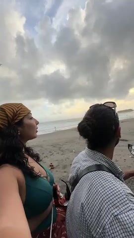 The video shows two people sitting on a motorcycle on a beach. The person in front is a man with dark hair pulled back in a bun. He wears a light colored, checkered button-down shirt. Strapped across his chest is a dark bag with light colored trim. Sunglasses are perched on top of his head. The person behind him is a woman with dark curly hair. She wears a green bikini top and a striped headscarf. Her right arm is visible and she holds a red patterned bag. The beach stretches out to the ocean under a cloudy, stormy sky.