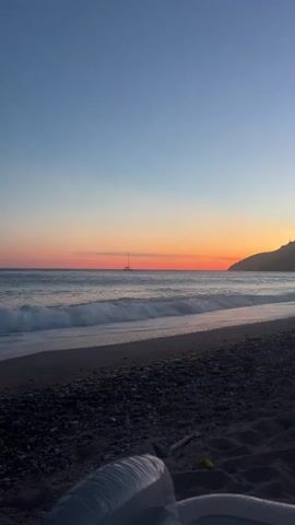 The video shows a tranquil beach scene at sunset. The sky transitions from a deep blue at the top to a vibrant orange and pink near the horizon, indicating the sun is setting. Gentle waves are rolling onto a dark sand and pebble beach. In the distance, a sailboat is visible on the calm sea. In the foreground, a portion of an inflated white object, possibly a pool float or mattress, is visible on the sand. The overall atmosphere is peaceful and serene, capturing the quiet beauty of a beach at dusk.