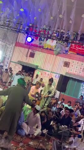 The video shows a crowded indoor gathering, possibly a religious event or celebration, with many men seated on the floor. The space is decorated with lights and fabric, creating a festive atmosphere. In the foreground, a person in a green outfit and headwear stands facing away from the camera, gesturing towards the crowd. Several individuals are visible in the background, some appearing to be performers or speakers, interacting with the audience. The lighting shifts throughout the clip, with various colors illuminating the scene. There are also musical instruments and sound equipment visible.