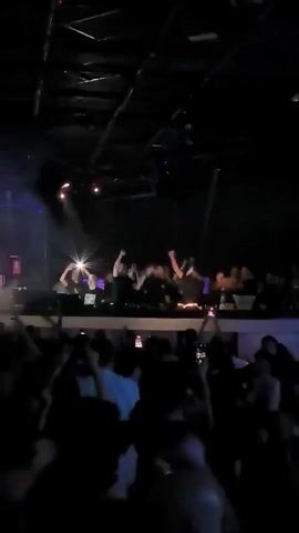A crowd of people with their hands in the air, dancing in a dimly lit, club-like atmosphere. There is a raised platform with a group of people on it, some of whom also have their hands up. There are flashing lights throughout the scene, sometimes obscuring details. Many people in the crowd appear to be recording with their phones.