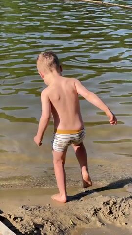 A shirtless boy in striped swim trunks stands at the edge of a body of water. The water has a green tint and light waves. The boy tentatively puts his foot in the water, then continues walking along the edge, looking out over the water.  The shoreline is sandy and there is a piece of wood on the left. The light is bright and casts a shadow of the boy on the ground.