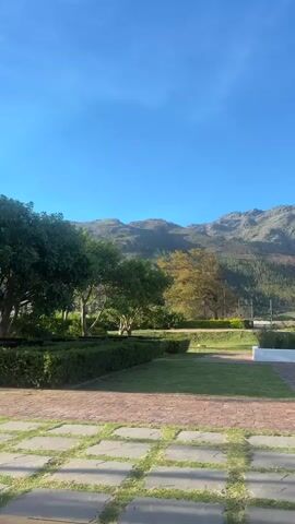 The video shows a landscape with a lawn and brick and stone pathway in the foreground, trees and shrubs in the middle ground, and mountains in the background under a clear blue sky. The scene appears to be a garden or park setting.