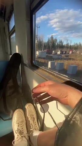A person is seen from a first-person perspective, sitting inside a moving train. Their legs, clad in white sneakers and socks, are propped up. In their hands, they are holding a pair of sunglasses. A black backpack, possibly containing a guitar due to its shape, rests on the seat next to them.

Through the train window, a bright, sunny daytime scene unfolds. The train is moving through a rural, forested landscape, characterized by numerous pine trees. The sky is partly cloudy with patches of clear blue. Power lines are visible running alongside the tracks. In the distance, some small houses or buildings can be glimpsed through the trees. The overall impression is one of a peaceful journey through the countryside, likely in an area like Susano, Russia, given the provided context. The sunlight casts shadows inside the train car, suggesting a time of day when the sun is relatively low, such as late afternoon.