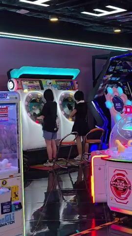In an arcade located in Shangcheng District, China, two individuals are actively engaged with a large, two-player rhythm game. The machine features prominent circular screens, each displaying graphics, and is illuminated by vibrant neon lights. One person, wearing a black t-shirt and blue shorts, stands facing their screen, while the other, in a black dress, is seated on a chair and also interacting with their respective screen.

Adjacent to the rhythm game, a claw machine is visible on the left, with plush toys displayed inside. To the right of the rhythm game, another arcade machine with a circular, illuminated design is present, displaying the text "BLUE SPACESHIP." The overall setting is a well-lit arcade with a glossy floor reflecting the colorful lights and machinery. The ambiance suggests a lively evening or indoor activity, with no indication of external weather conditions. The individuals appear focused on their gameplay.