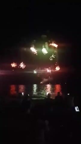 A night-time video shows a fireworks display over a body of water, with silhouettes of people watching from the shore in the foreground. The fireworks are multi-colored, with bursts of red, green, and white lights illuminating the dark sky. The reflections of the fireworks are visible on the water's surface, creating a shimmering effect. The scene suggests a celebratory event, possibly a festival or holiday.