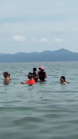 A group of people are standing in the ocean, with mountains in the background under a partly cloudy sky. Some individuals are partially submerged in the water, appearing to be enjoying themselves.