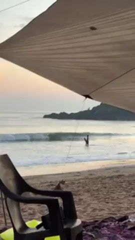 The video depicts a scenic beach scene, captured from under a makeshift shelter. The beach is sandy, and gentle waves are lapping at the shore. In the distance, a rocky landmass extends into the sea. A man with a beard and dark clothing is seen walking out of the water onto the beach, carrying a surfboard. A chair and blanket are visible in the foreground under the shelter, suggesting a relaxed or camping setting. The sky is pale with the light of dawn or dusk.