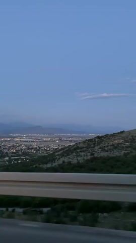 The video shows a view of a city at night from an elevated position, likely a roadside. The city is spread out with numerous lights visible. In the distance, mountains are silhouetted against the dusky blue sky. A roadside barrier is visible in the foreground, and some greenery can be seen on the side of the road. The camera seems to be moving, possibly from a car. The sky is a gradient of blue with some faint clouds.