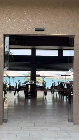 The video shows the entrance to a restaurant. The entrance is framed by a speckled wall with glass doors on either side. Through the entrance, the interior of the restaurant is visible. Tables and chairs are arranged on a tiled floor. Large windows offer a view of the ocean with umbrellas visible. People can be seen seated at some tables. The scene is bright with natural light. The overall impression is a calm and upscale setting.