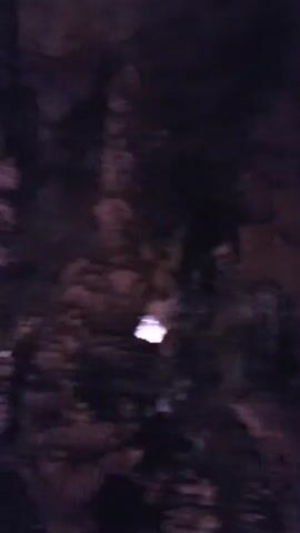 The video shows a dimly lit cave environment with rock formations. The camera is moving and panning, giving a shaky perspective of the cave's interior. There are several lights illuminating parts of the cave walls and ceiling. In the foreground, the silhouettes of people are visible, suggesting a tour group or visitors. The rock formations appear to be stalactites and stalagmites, typical of a cave. The lighting is predominantly in shades of purple and pink, creating an atmospheric effect. The overall impression is of a natural cave being explored by a group of people.
