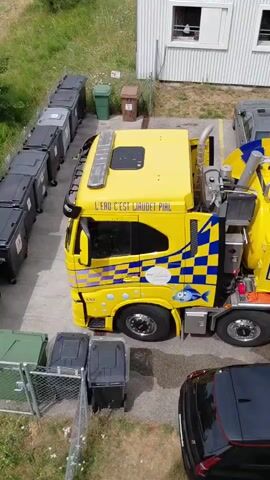 The video begins with a top-down view of a bright yellow vacuum truck parked next to a white building. Several large gray and green dumpsters are visible to the left of the truck, as well as some parked cars. The truck has the words "LPG C'EST L'URGENCE H24" printed on the side, along with a checkered pattern and a cartoon fish. A hose extends from the back of the truck towards a man in an orange uniform, who is operating a piece of equipment near a manhole cover. More cars are parked around the scene, and a street is visible in the background. The video is shot from an elevated perspective, likely from a drone or a higher floor of the building.