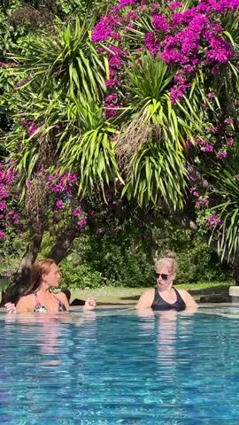 Two women are in an infinity pool, surrounded by lush vegetation. The pool's water is a vibrant blue, reflecting the sunlight. One woman has reddish hair and is wearing a colorful bikini top, while the other has blonde hair pulled back and is wearing a black swimsuit and sunglasses. They are facing each other and talking. Behind them, there's a dense collection of greenery including a tree with bright purple flowers. The setting seems tropical and peaceful.