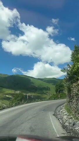 A POV video shows a car driving on a winding asphalt road through a mountainous, green landscape under a bright blue sky with fluffy white clouds. The road curves to the right, with a steep, rocky embankment on the right side covered in green foliage and trees. On the left side, there is a railing and a drop-off into a valley with more green hills. Road signs indicating a curve are visible. Sunlight illuminates the scene.