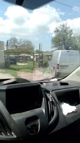 From the perspective of inside a vehicle, the video shows a front view of approaching a gated property on a partly cloudy day. The property is surrounded by a brick and metal fence, and a large, dark metal gate. A silver van is parked inside the gate. The vehicle moves slowly towards the gate. The dashboard of the vehicle is visible, including a screen, air vents, and the steering wheel.