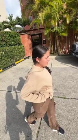 A young girl with dark hair pulled back in a ponytail is walking towards the camera. She is wearing a light tan oversized hoodie and brown cargo pants. She has a gold hoop earring in her left ear. The girl is in an outdoor setting, likely a driveway or a paved walkway. Behind her, a silver Toyota Tacoma pickup truck is parked. The truck has black rims. Lush green palm trees are visible in the background, as well as a brick building. The sun is shining, casting shadows on the ground, suggesting it is daytime. The girl appears to be smiling and looking at the camera. No text is visible in the video. The location context provided is Mérida, Venezuela.