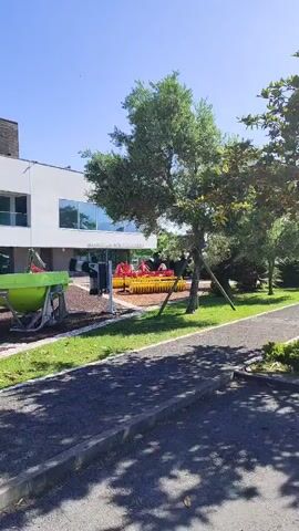 This video shows an outdoor display of agricultural equipment in front of a modern building. The building has a dark, slatted roof and a white facade with large, reflective windows. A sign on the building reads "RESTAURANTE GASTRONOMIA".

The agricultural equipment includes a bright red plow with yellow wheels and several other pieces of machinery in red, yellow, and green. These appear to be agricultural implements, possibly for plowing or tilling. They are displayed on a ground covered with dark gravel and mulch. In the foreground, there is a paved walkway and a grassy area. A tree with green foliage stands to the right of the frame, casting shadows on the ground. The sky is a clear, bright blue, suggesting a sunny day.

In the background, a white car is visible driving in front of the building. The overall scene appears to be a public space, possibly an exhibition or a park associated with the restaurant. The bright sunlight and clear sky indicate it is daytime. There are no people clearly visible in the frame, and the focus is on the agricultural machinery and the building. The presence of the equipment suggests a connection to local agriculture or a theme related to farming. The location appears to be outdoors, under a sunny sky, in what seems to be a well-maintained area.